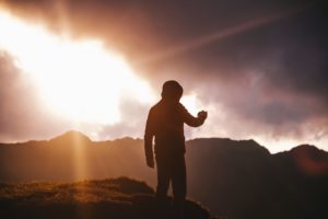 Man standing on mountain top with mountain ridge and sunrise in the background.