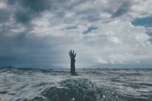 An arm and open hand reaching up out of choppy waters.  Backdrop of a stormy sky.